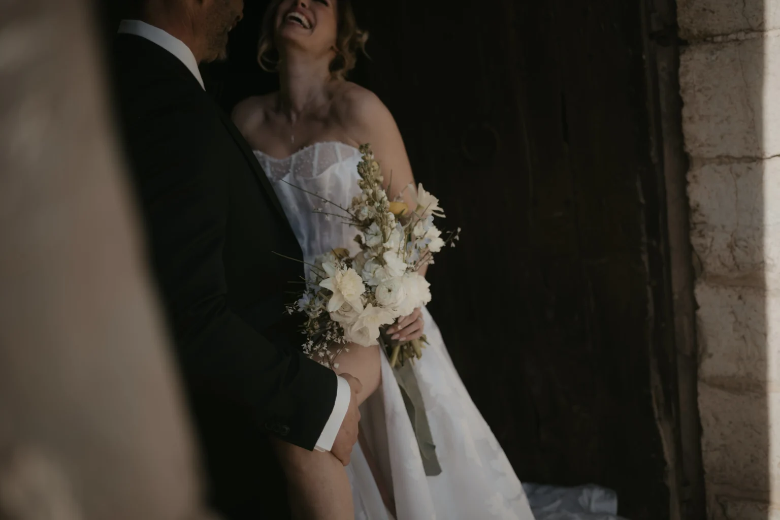 Brautpaar beim Fotoshooting. Der Bräutigam lehnt sich lässig an eine Hauswand und die Braut präsentiert ihren Brautstrauß.